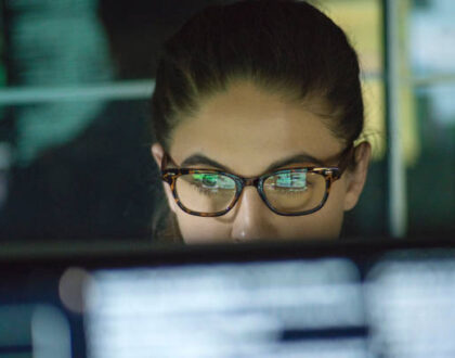 Close up stock photo of a young woman surrounded by monitors & their reflections displaying scrolling text & data.